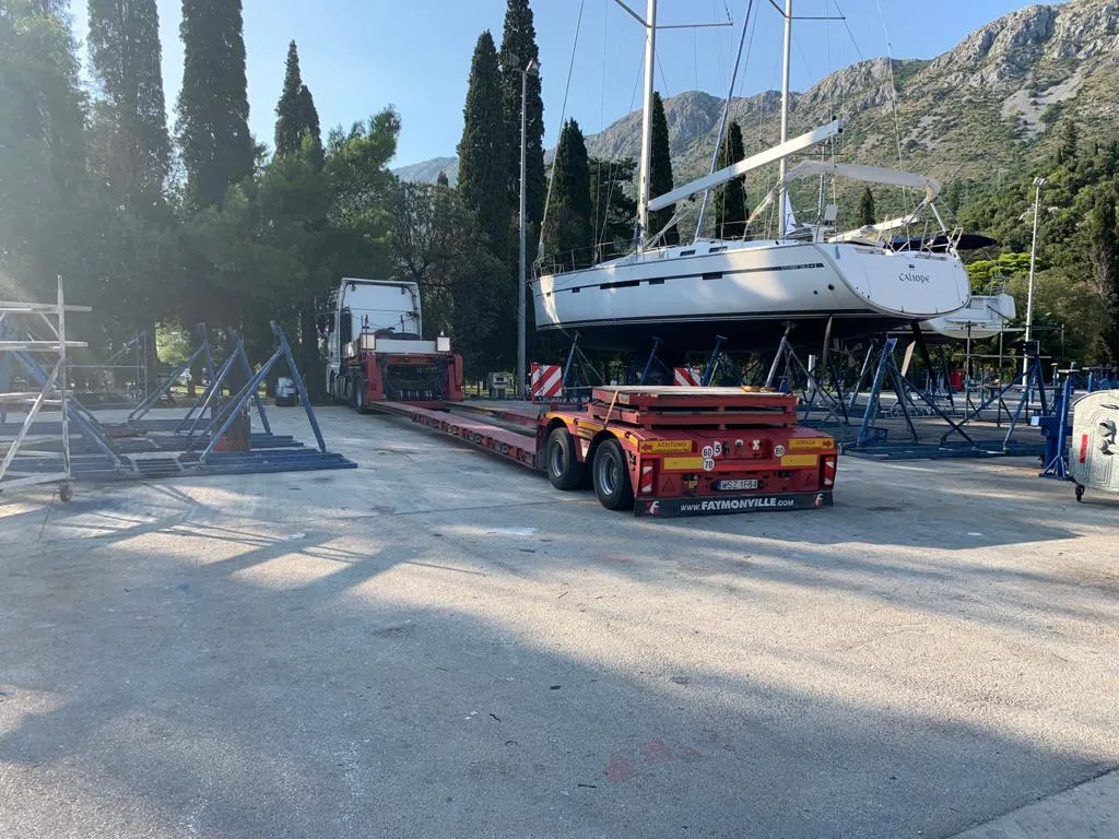 Sailing yacht on a red low-loader trailer in front of Adriatic mountains.