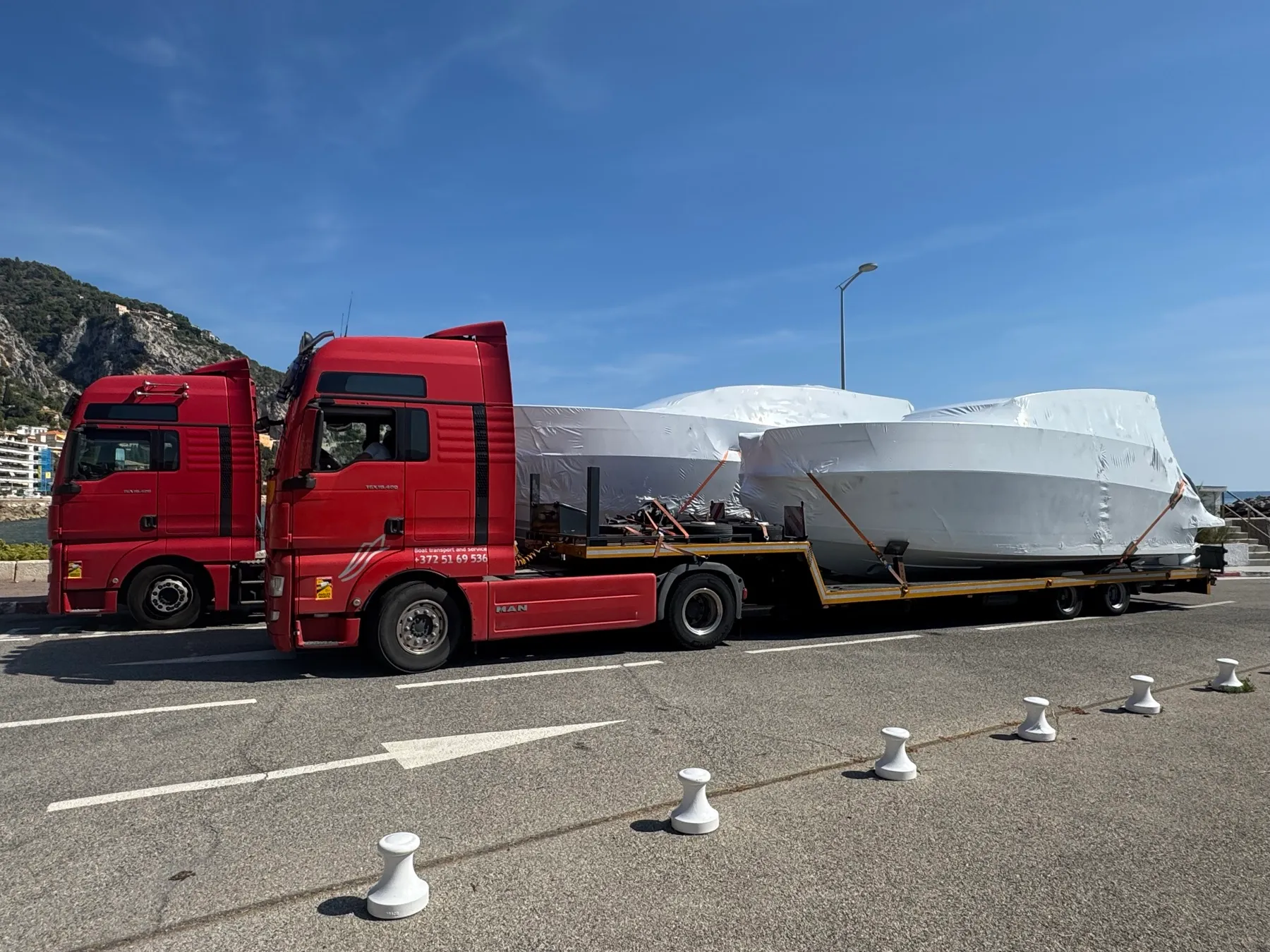 Two red trucks with shrink-wrapped cruisers on a Mediterranean coast road.