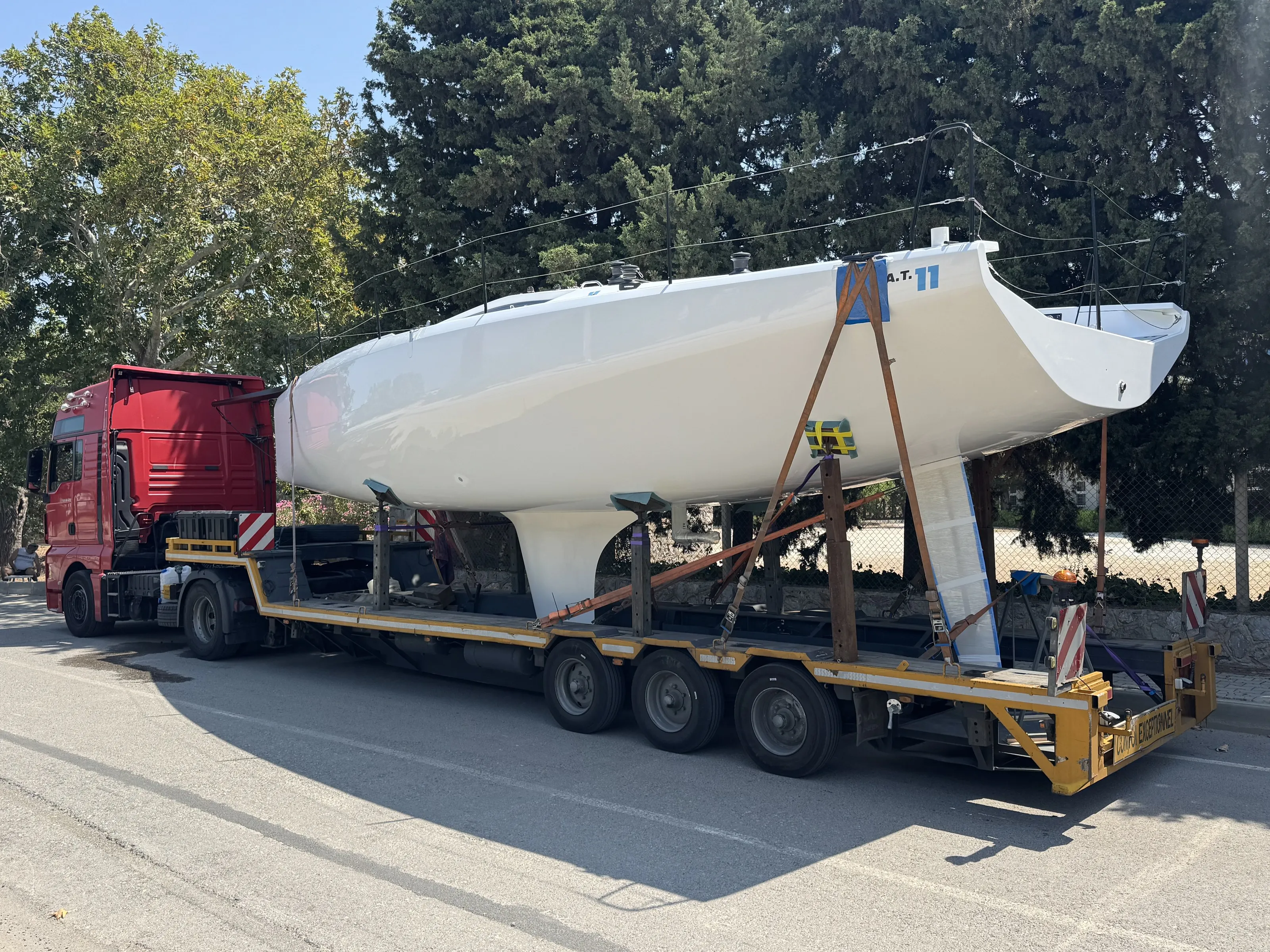 White sailing yacht secured on a yellow low-loader trailer behind a red MAN truck.